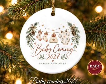 May include: A round white ceramic ornament featuring a clothesline illustration with two socks, a knit hat, and a baby onesie. The text reads Baby Coming 2027 and Sarah and Mike. It is decorated with pine branches, pinecones, and gold stars.