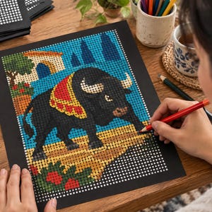 May include: A square coloring page featuring a black bull with a red and gold decorative blanket, standing on a tan ground against a blue background with a small building and trees, rendered in a grid-based pixel art style on black paper.