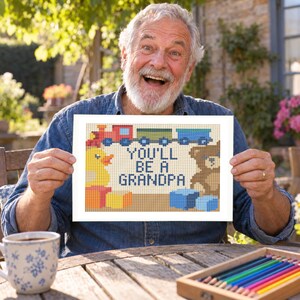 May include: A rectangular cross-stitch pattern featuring a red train, a yellow rubber duck, a brown teddy bear, and blue and yellow blocks. The text in the center reads YOU'LL BE A GRANDPA in dark blue capital letters against a light background.