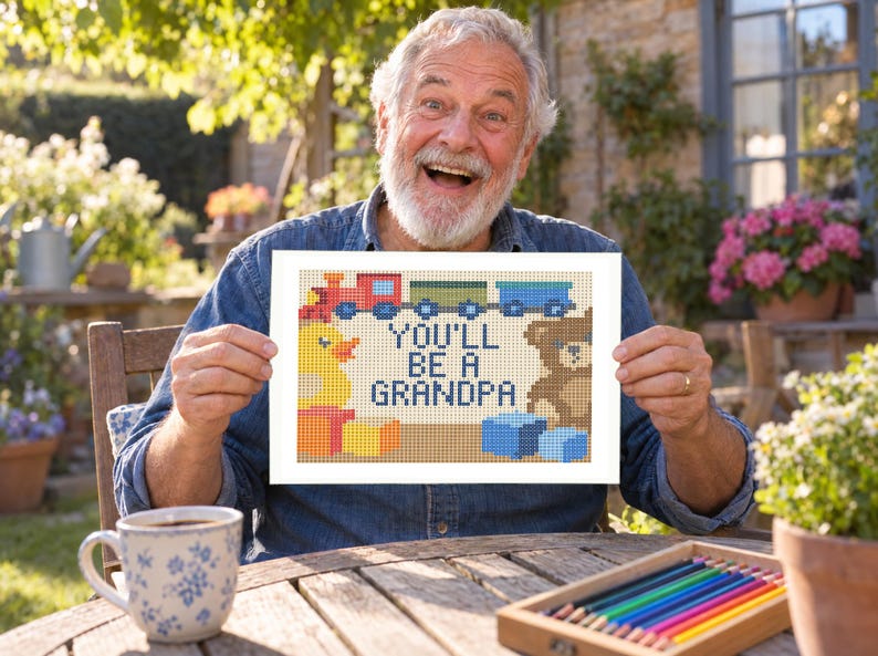 May include: A rectangular cross-stitch pattern featuring a red train, a yellow rubber duck, a brown teddy bear, and blue and yellow blocks. The text in the center reads YOU'LL BE A GRANDPA in dark blue capital letters against a light background.