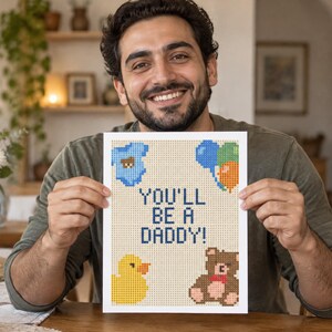 May include: A rectangular print featuring a pixelated design on a light beige background. The text reads YOU'LL BE A DADDY! in dark blue. Surrounding the text are pixelated illustrations of a blue baby onesie, colorful balloons, a yellow duck, and a brown teddy bear.