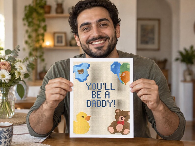 May include: A rectangular print featuring a pixelated design on a light beige background. The text reads YOU'LL BE A DADDY! in dark blue. Surrounding the text are pixelated illustrations of a blue baby onesie, colorful balloons, a yellow duck, and a brown teddy bear.