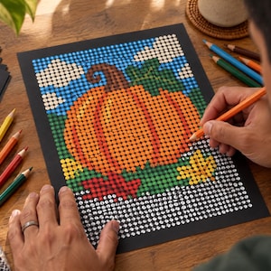 May include: A dot art project on black paper featuring a large orange pumpkin with a brown stem and green leaves against a blue sky with white clouds. The design is partially filled with colored dots, with some areas remaining as white circles.