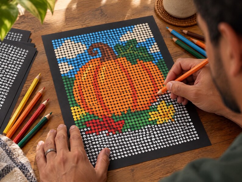 May include: A dot art project on black paper featuring a large orange pumpkin with a brown stem and green leaves against a blue sky with white clouds. The design is partially filled with colored dots, with some areas remaining as white circles.