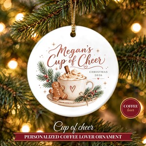Peut inclure: Une décoration ronde en céramique blanche ornée d'une tasse de café avec de la crème fouettée, un bâton de cannelle et un bonhomme en pain d'épices. Le texte indique Megan's Cup of Cheer et CHRISTMAS 2026. Elle est suspendue par un cordon doré.