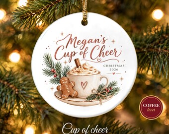 May include: A round white ceramic ornament featuring a graphic of a coffee cup with whipped cream, a cinnamon stick, and a gingerbread man. The text reads Megan's Cup of Cheer and CHRISTMAS 2026. It hangs from a gold cord.