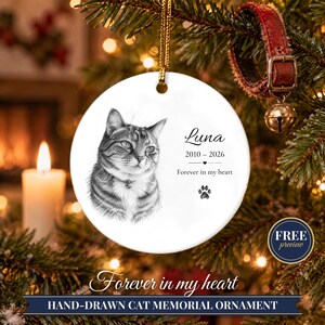 May include: A round white ceramic ornament featuring a black and white sketch of a cat. The text reads Luna, 2010-2026, and Forever in my heart, accompanied by a small heart icon and a paw print graphic. It hangs from a gold cord.