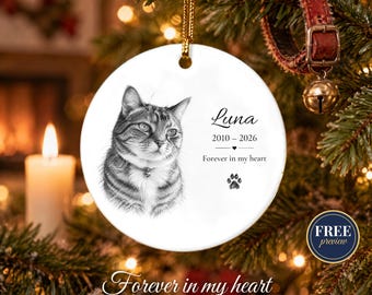 May include: A round white ceramic ornament featuring a black and white sketch of a cat. The text reads Luna, 2010-2026, and Forever in my heart, accompanied by a small heart icon and a paw print graphic. It hangs from a gold cord.