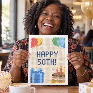 May include: A rectangular white sign featuring a pixelated design with the text HAPPY 50TH! in dark blue. The graphic includes colorful balloons in red, yellow, and blue, a blue gift box, and a brown birthday cake with three lit candles.