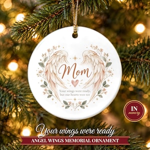May include: A round white ceramic memorial ornament featuring a pair of light brown angel wings, a small heart, and floral accents. The text reads Mom and Your wings were ready, but our hearts were not. It hangs from a gold cord.