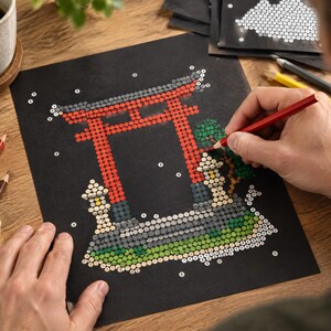 以下が含まれることがあります： 黒い紙に描かれた数字で塗るアートプロジェクトで、赤い鳥居、石灯籠、緑の葉が描かれています。デザインは小さな番号付きの円で構成されており、赤い鉛筆で鳥居の一部を塗りつぶしている様子です。
