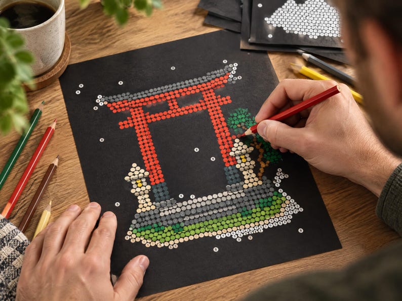 以下が含まれることがあります： 黒い紙に描かれた数字で塗るアートプロジェクトで、赤い鳥居、石灯籠、緑の葉が描かれています。デザインは小さな番号付きの円で構成されており、赤い鉛筆で鳥居の一部を塗りつぶしている様子です。