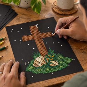 May include: A black sheet of paper featuring a dot-to-dot drawing of a brown cross set atop a green grassy mound with small rocks. The artwork is composed of numerous small circles, and a brown pencil is being used to connect the dots on the paper.