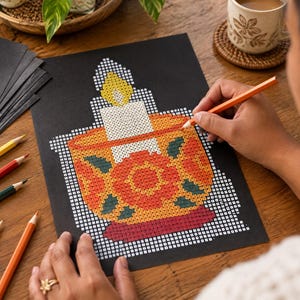 May include: A black paper sheet featuring a grid-based pixel art design of a lit candle in a decorative bowl. The candle flame is yellow, the candle is white, and the bowl displays orange, red, and dark green floral patterns on a yellow base.