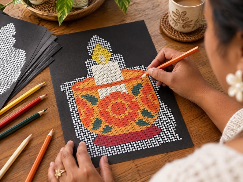May include: A black paper sheet featuring a grid-based pixel art design of a lit candle in a decorative bowl. The candle flame is yellow, the candle is white, and the bowl displays orange, red, and dark green floral patterns on a yellow base.