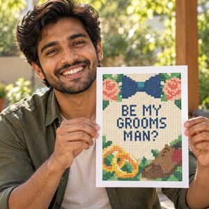 May include: A rectangular print featuring a cross-stitch style design with the text BE MY GROOMSMAN? in dark blue. The design includes a blue bow tie, two pink roses, two gold rings, and a brown dress shoe, all set against a white background.