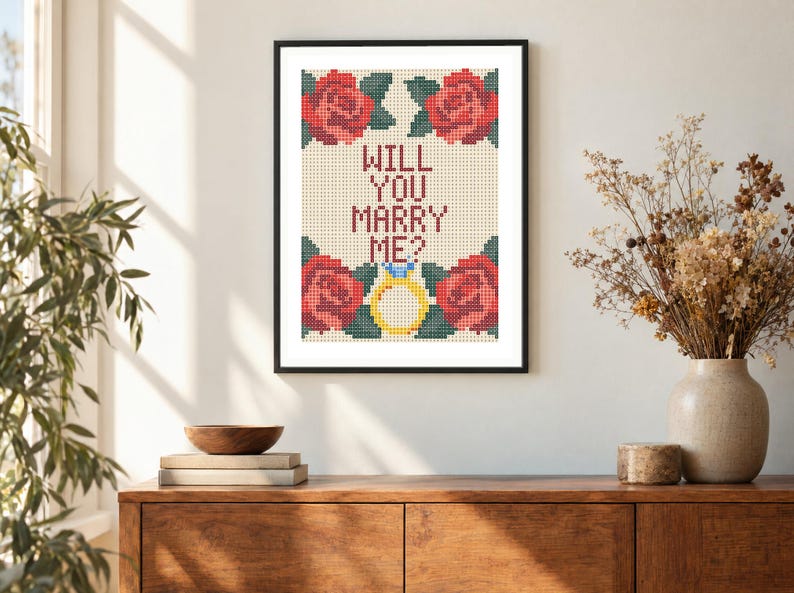 May include: A framed cross-stitch embroidery featuring the text WILL YOU MARRY ME? in dark red letters, surrounded by four red roses with green leaves and a yellow engagement ring with a blue gemstone, all set against a cream-colored grid.