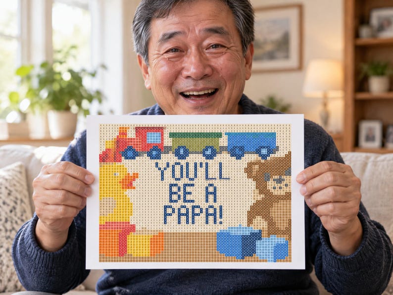 May include: A rectangular print featuring a pixelated design with the text YOU'LL BE A PAPA! in blue. The image includes a red and green train, a yellow rubber duck, a brown teddy bear, and blue blocks, all rendered in a grid-like pattern.
