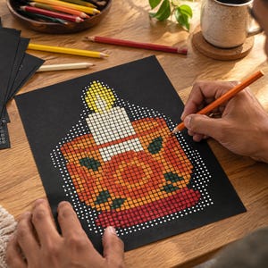 May include: A black sheet of paper featuring a grid-based pixel art design of a lit candle in a holder. The candle has a yellow flame, a white body, and sits in an orange and red base with dark green accents. The design is composed of small squares.