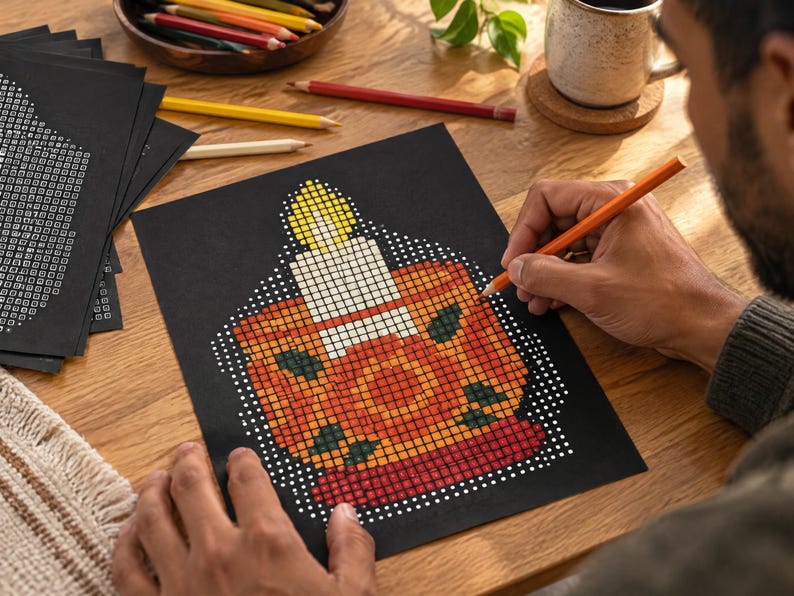 May include: A black sheet of paper featuring a grid-based pixel art design of a lit candle in a holder. The candle has a yellow flame, a white body, and sits in an orange and red base with dark green accents. The design is composed of small squares.