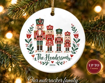 May include: A round white ceramic ornament featuring a graphic of four nutcracker figures in red and green uniforms. Below the figures are the names Dad, Mom, Sam, and Zoe. The text The Hendersons and 2026 are printed in the center.