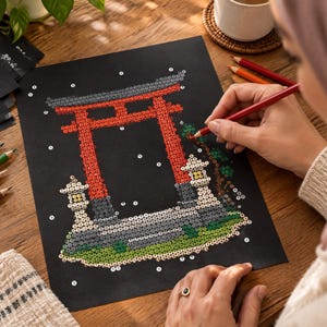 Pu&ograve; includere: Un foglio di carta nero con un disegno puntinato di un torii rosso e lanterne di pietra. Il motivo &egrave; parzialmente riempito con cerchi colorati. Una matita rossa viene utilizzata per unire i punti sul foglio appoggiato su una superficie in legno.