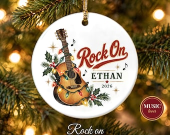 May include: A round white ceramic Christmas ornament featuring an acoustic guitar wrapped in colorful holiday lights, holly sprigs, and musical notes. The text reads Rock On, ETHAN, and 2026 in red and dark green lettering with gold star accents.
