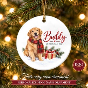May include: A round white ceramic Christmas ornament featuring a golden retriever wearing a red and green plaid scarf. The text reads Buddy, Christmas 2026, with a small red heart and a gift box with a red bow on a snowy surface.