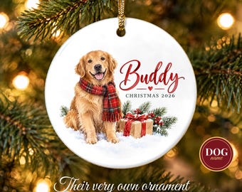 May include: A round white ceramic Christmas ornament featuring a golden retriever wearing a red and green plaid scarf. The text reads Buddy, Christmas 2026, with a small red heart and a gift box with a red bow on a snowy surface.