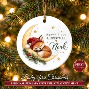 May include: A round white ceramic ornament featuring a sleeping fox wearing a red knit hat, resting on a crescent moon. The text reads Baby's First Christmas, Noah, and 2026. It includes gold stars, pine branches, and red berries, suspended by a gold cord.