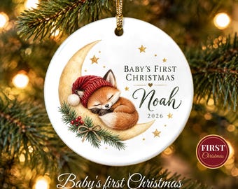 May include: A round white ceramic ornament featuring a sleeping fox wearing a red knit hat, resting on a crescent moon. The text reads Baby's First Christmas, Noah, and 2026. It includes gold stars, pine branches, and red berries, suspended by a gold cord.