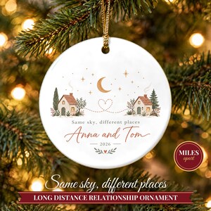 May include: A round white ceramic ornament featuring two illustrated houses connected by a dotted heart line under a crescent moon and stars. The text reads Same sky, different places, Anna and Tom, and 2026, with a gold hanging cord.