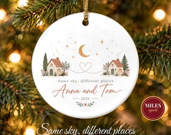 May include: A round white ceramic ornament featuring two illustrated houses connected by a dotted heart line under a crescent moon and stars. The text reads Same sky, different places, Anna and Tom, and 2026, with a gold hanging cord.