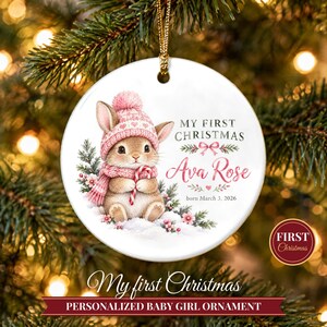 May include: A round white ceramic ornament featuring a brown bunny wearing a pink hat and scarf, holding a candy cane. The text reads My First Christmas, Ava Rose, and born March 3, 2026, with pink floral accents and a gold hanging cord.