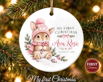 May include: A round white ceramic ornament featuring a brown bunny wearing a pink hat and scarf, holding a candy cane. The text reads My First Christmas, Ava Rose, and born March 3, 2026, with pink floral accents and a gold hanging cord.