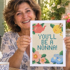 May include: A rectangular white card featuring a pixelated cross-stitch design with the text YOU'LL BE A NONNA! in blue. The design includes a red teapot, a yellow lemon, pink flowers, and blue flowers arranged around the central text.