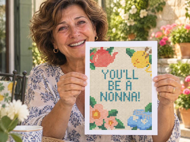 May include: A rectangular white card featuring a pixelated cross-stitch design with the text YOU'LL BE A NONNA! in blue. The design includes a red teapot, a yellow lemon, pink flowers, and blue flowers arranged around the central text.