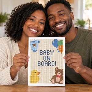 May include: A rectangular sign featuring a pixel art design on a light beige background. The text reads BABY ON BOARD! in dark blue. The design includes a blue baby onesie, a cluster of blue, green, and orange balloons, a yellow rubber duck, and a brown teddy bear.