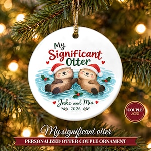 Puede incluir: Un adorno navideño redondo de cerámica blanca con dos nutrias marrones con gorros de Papá Noel flotando en agua azul. El texto dice My Significant Otter, Jake and Mia, y 2026, decorado con corazones rojos y hojas de acebo verdes.