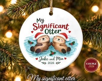 May include: A round white ceramic Christmas ornament featuring two brown otters wearing red Santa hats floating in blue water. The text reads My Significant Otter, Jake and Mia, and 2026, accented with red hearts and green holly leaves.