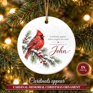 May include: A round white ceramic Christmas ornament featuring a red cardinal perched on a snowy pine branch with red berries. The text reads: Cardinals appear when angels are near, In loving memory of John. It hangs from a gold cord.