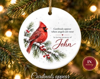 Puede incluir: Un adorno navideño redondo de cerámica blanca con un cardenal rojo sobre una rama de pino nevada con bayas rojas. El texto dice: Cardinals appear when angels are near, In loving memory of John. Incluye un cordón dorado.