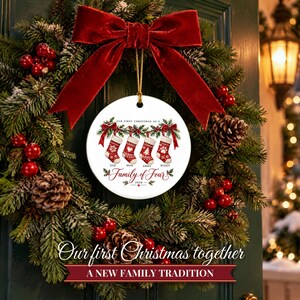 May include: A round white ceramic ornament featuring a graphic of four red and white Christmas stockings hanging from a garland. The text reads Our First Christmas as a Family of Four 2026, with names Dad, Mom, Emma, and Buddy below each stocking.
