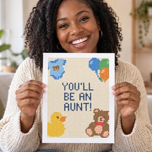 May include: A rectangular white card featuring a pixel-art style design with the text YOU'LL BE AN AUNT! in dark blue. The card includes a light blue baby onesie, a cluster of blue, green, and orange balloons, a yellow rubber duck, and a brown teddy bear.
