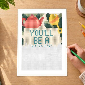 May include: A cross-stitch pattern printed on white paper featuring a red teapot, a yellow lemon, and green leaves at the top. The text "YOU'LL BE A" is printed in blue block letters in the center, with a grid of empty squares below.