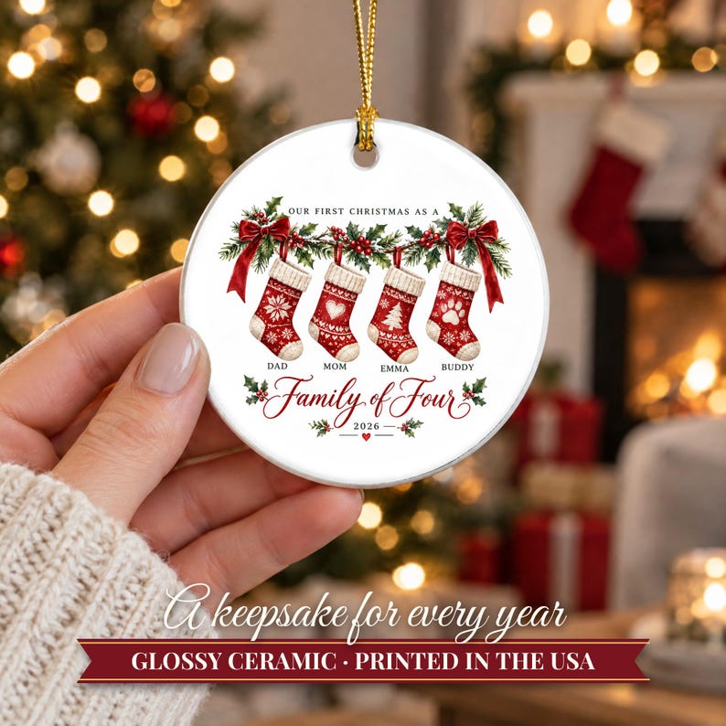 May include: A round white ceramic Christmas ornament featuring a printed illustration of four red and white stockings hanging from a green holly garland with red bows. The text reads Our First Christmas as a Family of Four, 2026, Dad, Mom, Emma, Buddy.