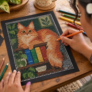 Puede incluir: Una hoja de arte estilo pintura por n&uacute;meros sobre papel negro que muestra un gato naranja esponjoso descansando en una estanter&iacute;a llena de libros coloridos. La imagen se compone de una cuadr&iacute;cula de peque&ntilde;os c&iacute;rculos numerados y una mano colorea.