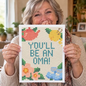 Puede incluir: Un lienzo rectangular de diamond painting con un patrón de cuadrícula y el texto YOU'LL BE AN OMA! en azul. El diseño incluye una tetera roja, un limón amarillo y flores rosas y azules, compuestas por pequeños cuadrados con códigos.
