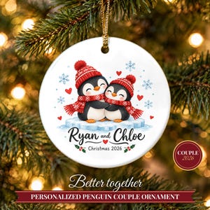 May include: A round white ceramic ornament featuring two penguins wearing red knit hats and scarves, hugging on an ice floe. The text reads Ryan and Chloe, Christmas 2026, with red hearts, blue snowflakes, and green holly accents. It hangs from a gold cord.
