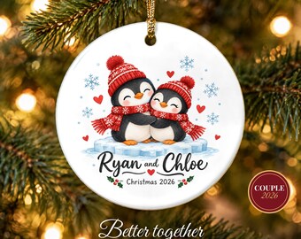 May include: A round white ceramic ornament featuring two penguins wearing red knit hats and scarves, hugging on an ice floe. The text reads Ryan and Chloe, Christmas 2026, with red hearts, blue snowflakes, and green holly accents. It hangs from a gold cord.